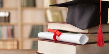 Imagem de um diploma enrolado com fita vermelha e um capelo de formatura, sobre uma pilha de livros