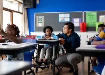 Professor e aluno em cadeira de rodas interagem em sala de aula inclusiva, com outros alunos ao fundo.