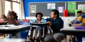 Professor e aluno em cadeira de rodas interagem em sala de aula inclusiva, com outros alunos ao fundo.