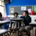 Professor e aluno em cadeira de rodas interagem em sala de aula inclusiva, com outros alunos ao fundo.