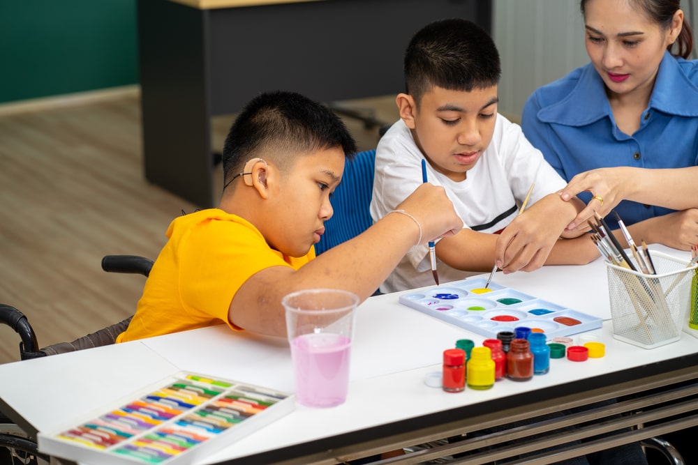Professor orientando dois alunos, um deles em cadeira de rodas e com aparelho auditivo, durante atividade de pintura em sala de aula inclusiva. Mostra o porque a educação inclusiva importa. 