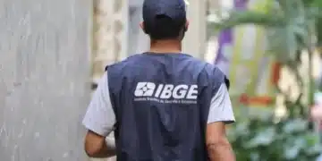 Trabalhador do IBGE usando colete azul, caminhando por uma rua estreita.