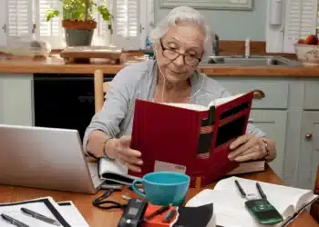 Idosa estudando em casa com livro e laptop sobre a mesa.