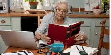 Idosa estudando em casa com livro e laptop sobre a mesa.