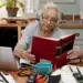 Idosa estudando em casa com livro e laptop sobre a mesa.