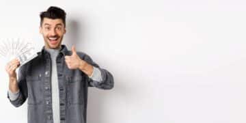 Homem sorridente segurando notas de dinheiro e fazendo sinal de positivo.