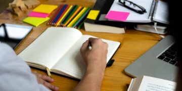 Pessoa escrevendo em um caderno com material de apoio, como lápis coloridos, post-its e livros, representando um ambiente de estudos.