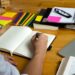 Pessoa escrevendo em um caderno com material de apoio, como lápis coloridos, post-its e livros, representando um ambiente de estudos.