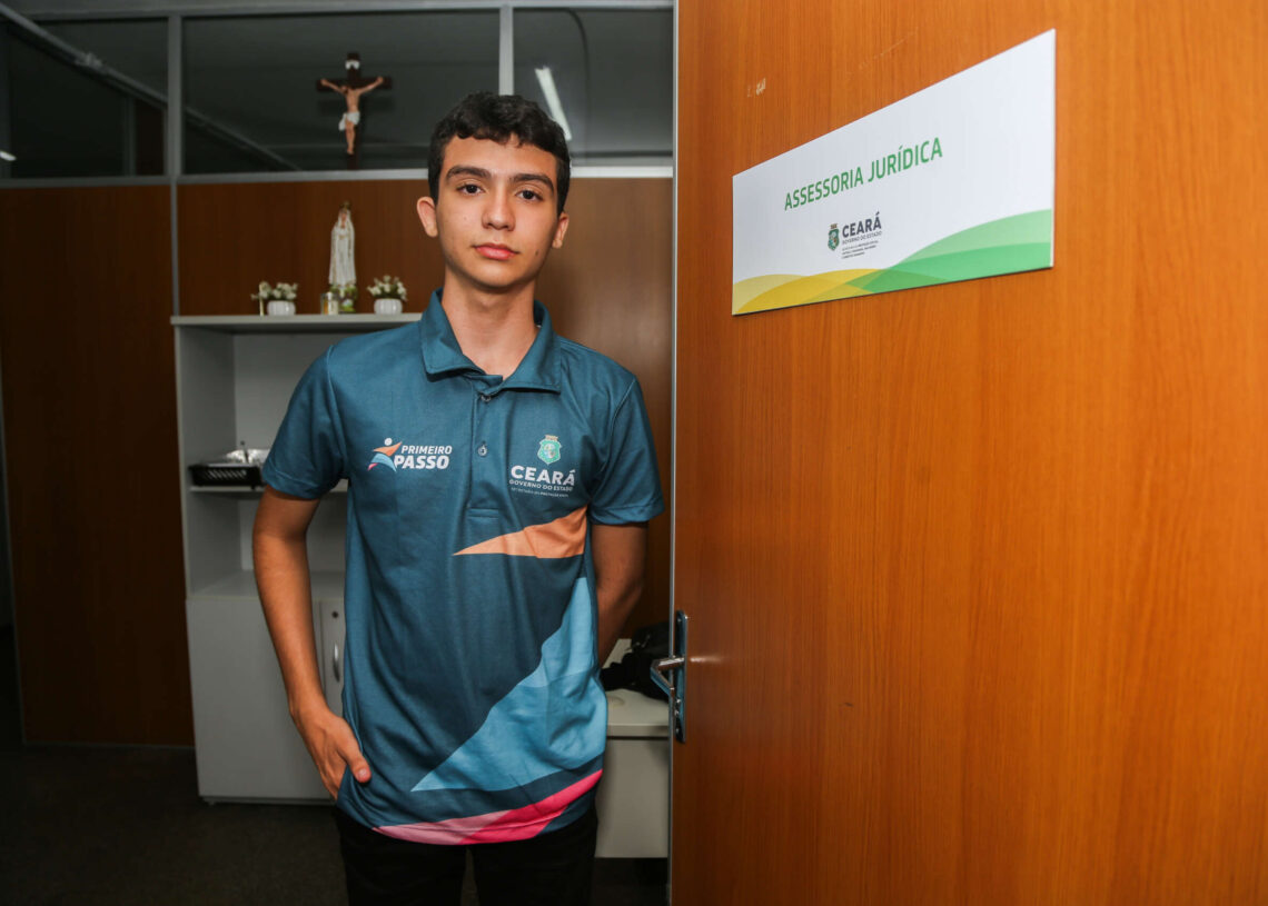 Jovem participando do programa Primeiro Passo, em ambiente corporativo, ao lado da placa de Assessoria Jurídica.