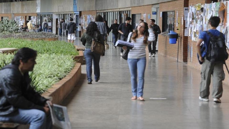 Corredor interno de Universidade, com estudantes circulando e painéis de avisos nas paredes.