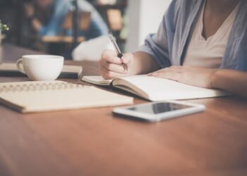 Imagem de uma pessoa escrevendo em um caderno com xícara de café e celular na mesa, simbolizando um momento de reflexão sobre a dúvida entre a escrita "amar o próximo" e "amar ao próximo".