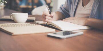 Imagem de uma pessoa escrevendo em um caderno com xícara de café e celular na mesa, simbolizando um momento de reflexão sobre a dúvida entre a escrita "amar o próximo" e "amar ao próximo".