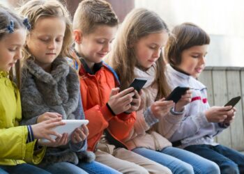 Grupo de crianças sentadas, concentradas em seus smartphones e tablets, ilustrando o uso crescente da tecnologia na infância e o debate sobre o "PL da Adultização".