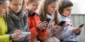 Grupo de crianças sentadas, concentradas em seus smartphones e tablets, ilustrando o uso crescente da tecnologia na infância e o debate sobre o "PL da Adultização".