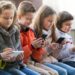 Grupo de crianças sentadas, concentradas em seus smartphones e tablets, ilustrando o uso crescente da tecnologia na infância e o debate sobre o "PL da Adultização".