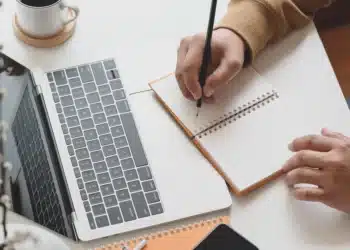 Uma pessoa escrevendo em um caderno ao lado de um notebook e estudando sobre o novo acordo ortográfico.