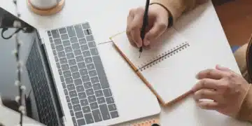 Uma pessoa escrevendo em um caderno ao lado de um notebook e estudando sobre o novo acordo ortográfico.