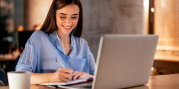 Mulher sorridente estudando com fones de ouvido, escrevendo em um caderno enquanto utiliza um notebook, representado as pessoas que escolhem cursos online.