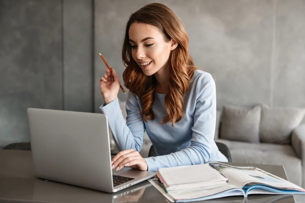 Mulher sorrindo enquanto estuda no computador, com livros e anotações ao lado, representando o estudo em casa