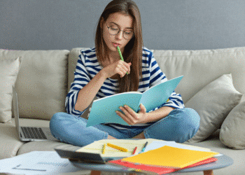 ulher jovem em casa, sentada no sofá, estudando as classes de palavras com um livro e notebook ao lado.
