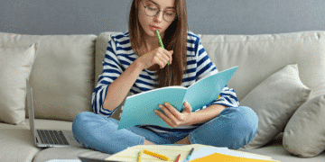 ulher jovem em casa, sentada no sofá, estudando as classes de palavras com um livro e notebook ao lado.