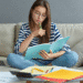 ulher jovem em casa, sentada no sofá, estudando as classes de palavras com um livro e notebook ao lado.