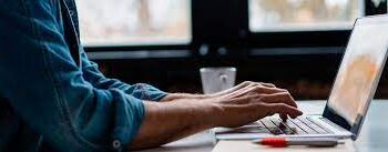 Pessoa digitando no computador, com foco nas mãos e teclado, trabalhando em um ambiente de escritório moderno.