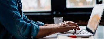 Pessoa digitando no computador, com foco nas mãos e teclado, trabalhando em um ambiente de escritório moderno.