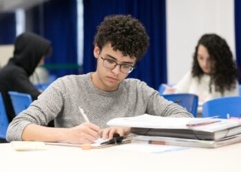 Jovem estudando em ambiente com outros estudantes, focado em anotações em mesa com materiais de concordância verbal e nominal.