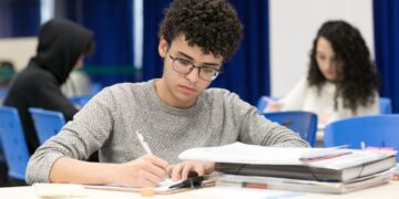 Jovem estudando em ambiente com outros estudantes, focado em anotações em mesa com materiais de concordância verbal e nominal.
