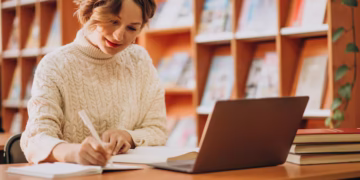 Mulher jovem e sorridente escrevendo em um caderno enquanto usa um notebook em uma biblioteca. Reflexão sobre o novo acordo ortográfico.