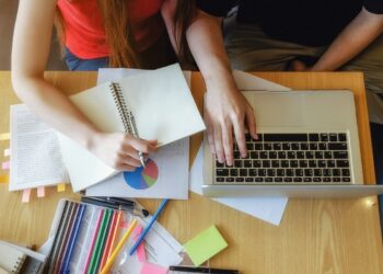 Mãos de uma pessoa escrevendo em um caderno enquanto outra pessoa digita no computador, com diversos materiais sobre estudo gramatical da concordância nominal de escritório sobre a mesa,