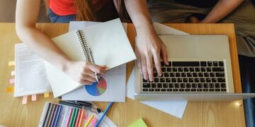 Mãos de uma pessoa escrevendo em um caderno enquanto outra pessoa digita no computador, com diversos materiais sobre estudo gramatical da concordância nominal de escritório sobre a mesa,
