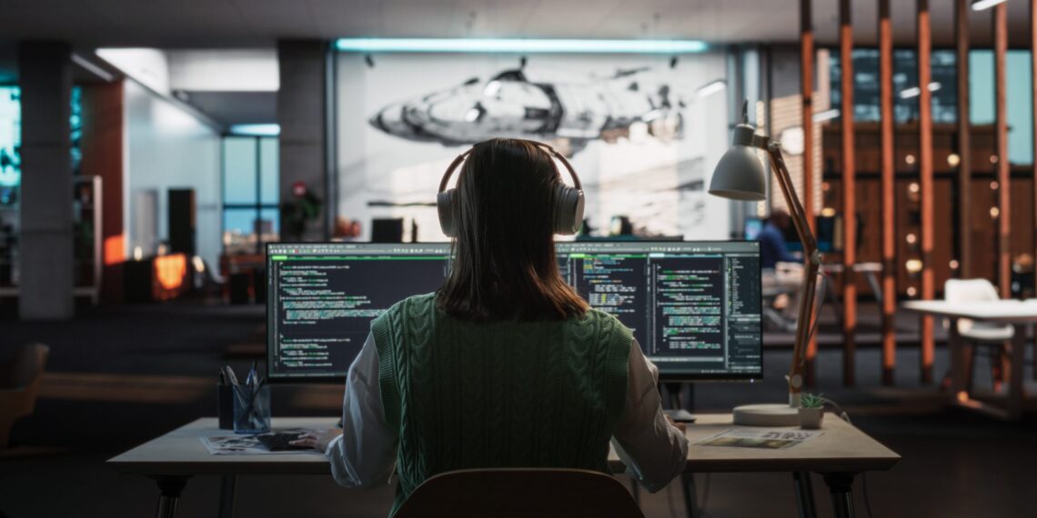 Pessoa programando em dois monitores, com código de computador visível, usando fones de ouvido, em um ambiente moderno de escritório.