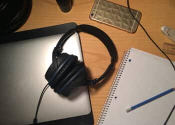 Fones de ouvido e outros itens sobre uma mesa de trabalho, incluindo um notebook, celular e caderno, referenciando-se aos estudos para vestibular com música.