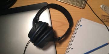 Fones de ouvido e outros itens sobre uma mesa de trabalho, incluindo um notebook, celular e caderno, referenciando-se aos estudos para vestibular com música.