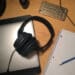Fones de ouvido e outros itens sobre uma mesa de trabalho, incluindo um notebook, celular e caderno, referenciando-se aos estudos para vestibular com música.