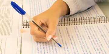 Mão de estudante escrevendo em um caderno com caneta azul, ao lado de livros e anotações, simbolizando a preparação para redação do Enem.