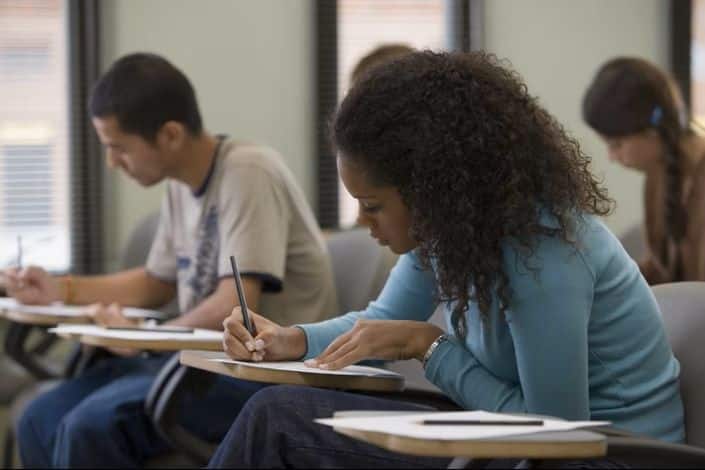 Estudantes fazendo a prova em ambiente de vestibular, com foco em uma mulher escrevendo."