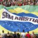 Multidão reunida em protesto com bandeira gigante do Brasil e faixa “Sem Anistia” ao centro.