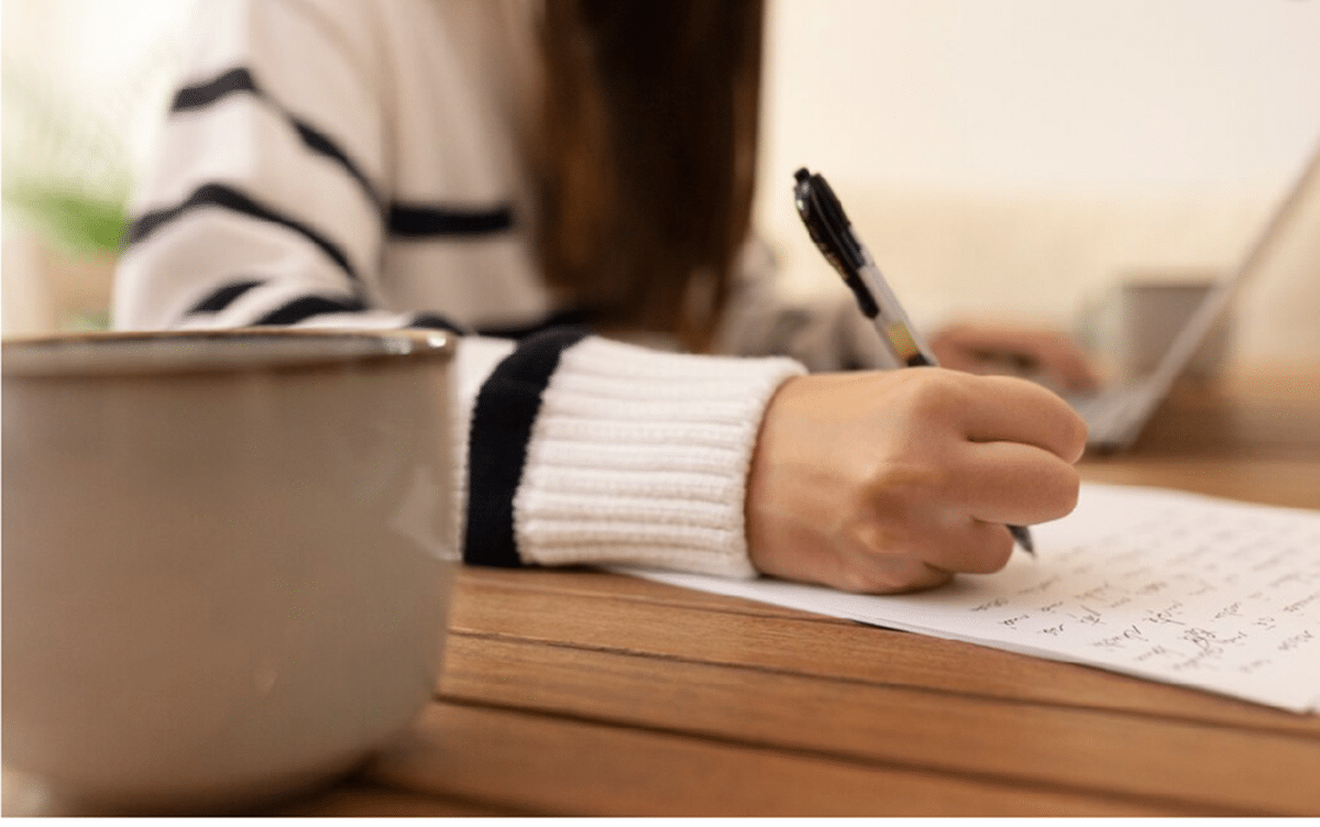 Estudante escrevendo em papel com caneta ao lado de xícara de café em mesa de madeira