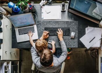 Visão superior de um pai ou mãe trabalhando em casa, com um notebook e um filho sentado no colo ajudando a digitar no teclado.