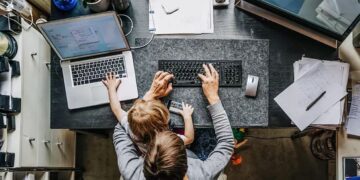 Visão superior de um pai ou mãe trabalhando em casa, com um notebook e um filho sentado no colo ajudando a digitar no teclado.