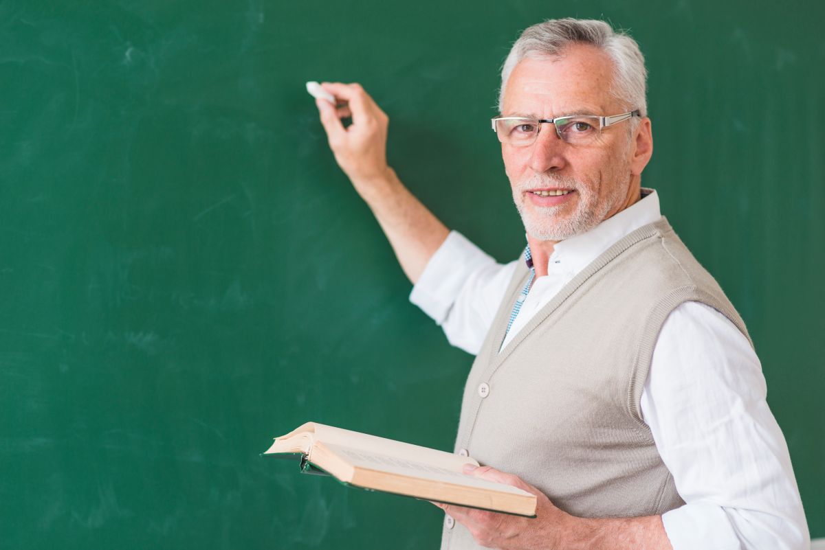 Professor mais velho sorri enquanto escreve na lousa, ilustrando o profissional de educação e a nova carteira digital.