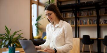 ovem mulher com óculos e suéter branco, sorrindo enquanto trabalha em um notebook em uma mesa de casa, representado o home office.