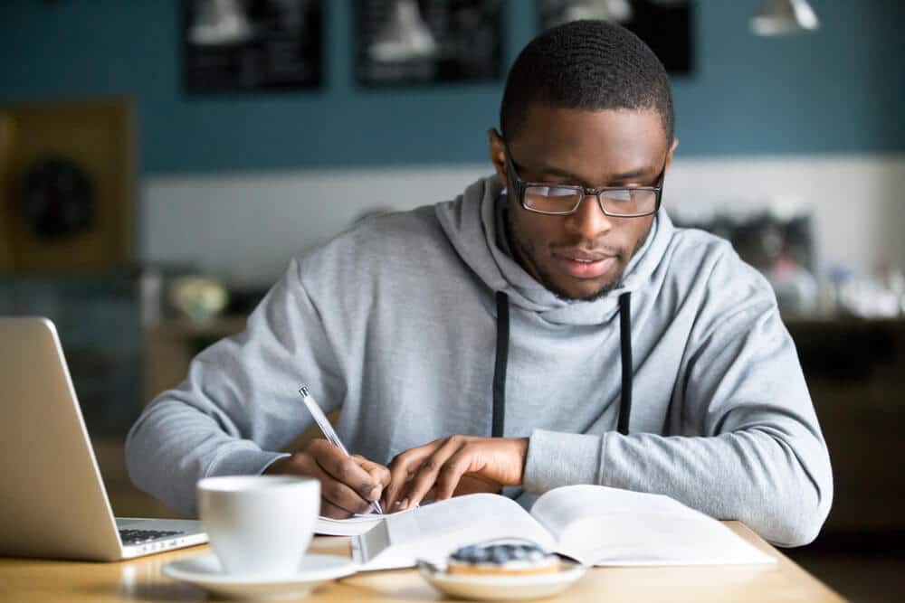 Homem estudando física em um café com notebook, anotando em livro aberto, com foco em sua preparação para exames.