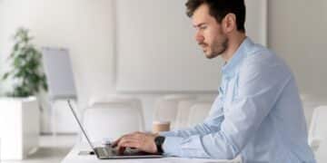 Homem jovem em ambiente de escritório, trabalhando em seu notebook. Representa um profissional em home office, um dos temas centrais no debate sobre monitoramento e produtividade
