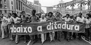 Manifestação histórica com faixas de protesto contra a ditadura militar, simbolizando a resistência popular e a luta pela democracia, referenciando aos golpes de estado que são cobrados em provas.