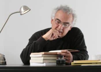 Imagem de Milton Hatoum lendo um livro, com pilha de livros sobre a mesa e uma luminária de mesa, autor que é muito citado no Enem e vestibulares.