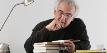 Imagem de Milton Hatoum lendo um livro, com pilha de livros sobre a mesa e uma luminária de mesa, autor que é muito citado no Enem e vestibulares.